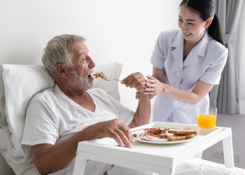 senior man with smiling nurse, takes care breakfast and discussion and cheer on bed at nursing home