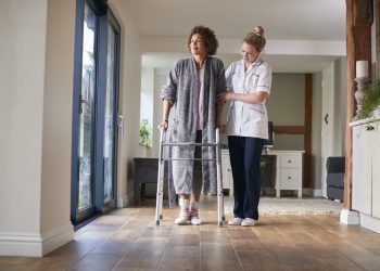 Mature Woman In Dressing Gown Using Walking Frame Being Helped By Female Nurse
